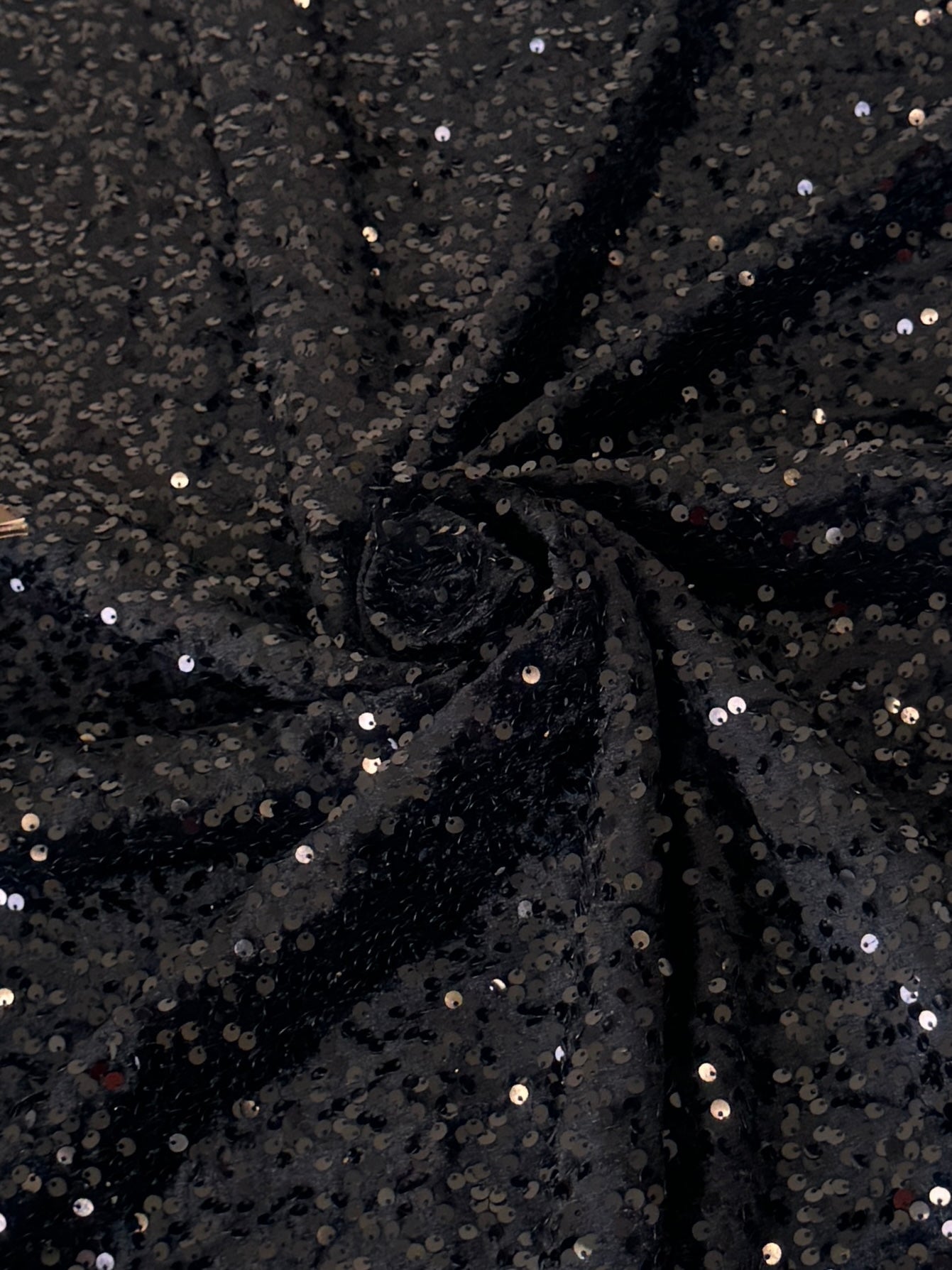 Black sequined fabric draped in a spiral pattern, showing shimmering silver and gold reflective sequins on a dark background.